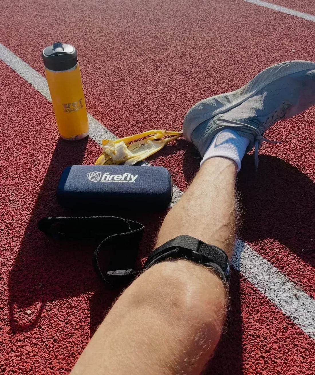 Athlete relaxing on a running track after training while wearing a Firefly recovery device, with a water bottle and banana nearby.
