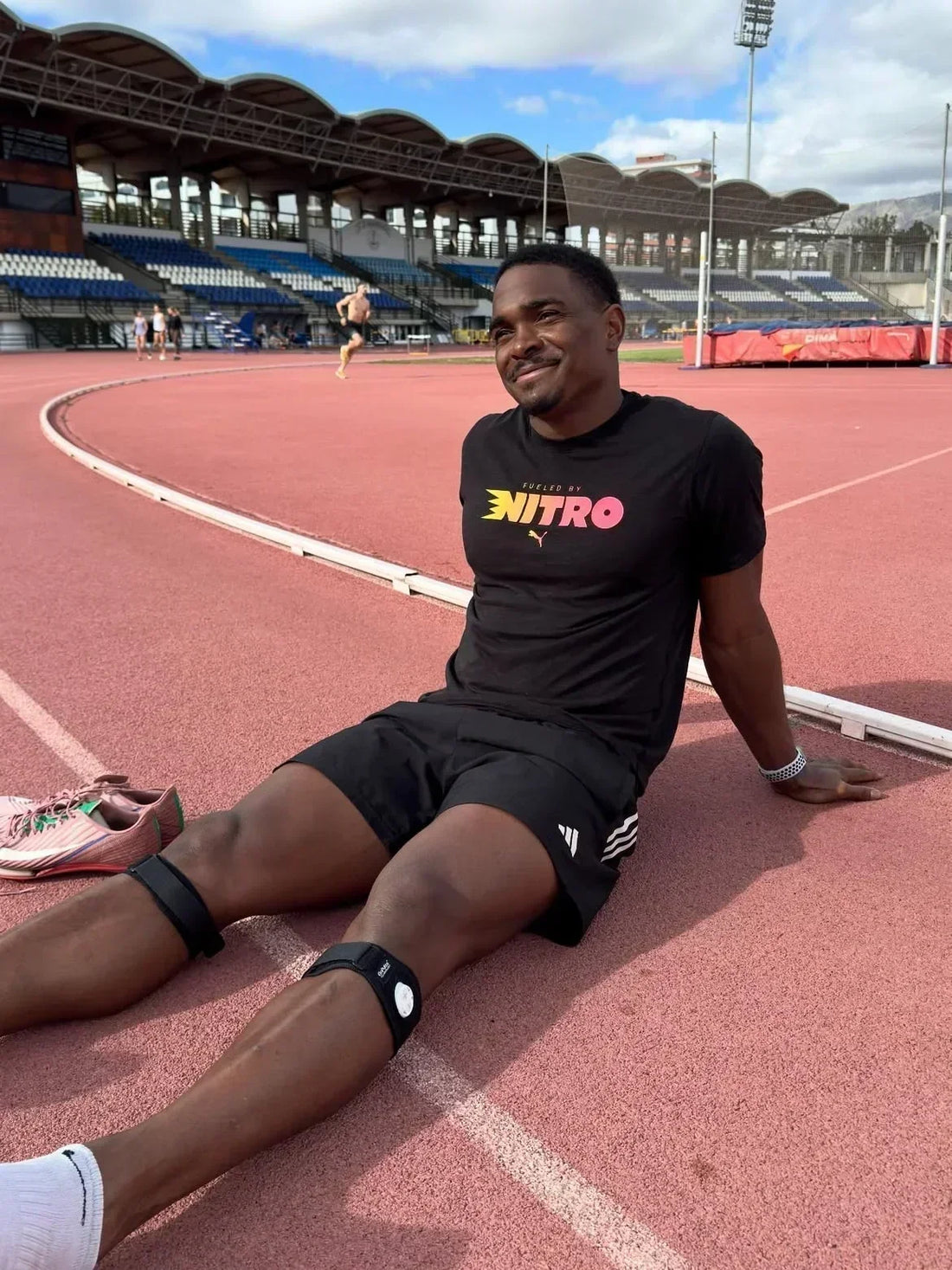 Runner wearing a black T-shirt and shorts sits beside a running track using the Firefly Recovery device after a 5K training session. The athlete is taking time to recover following an outdoor run.