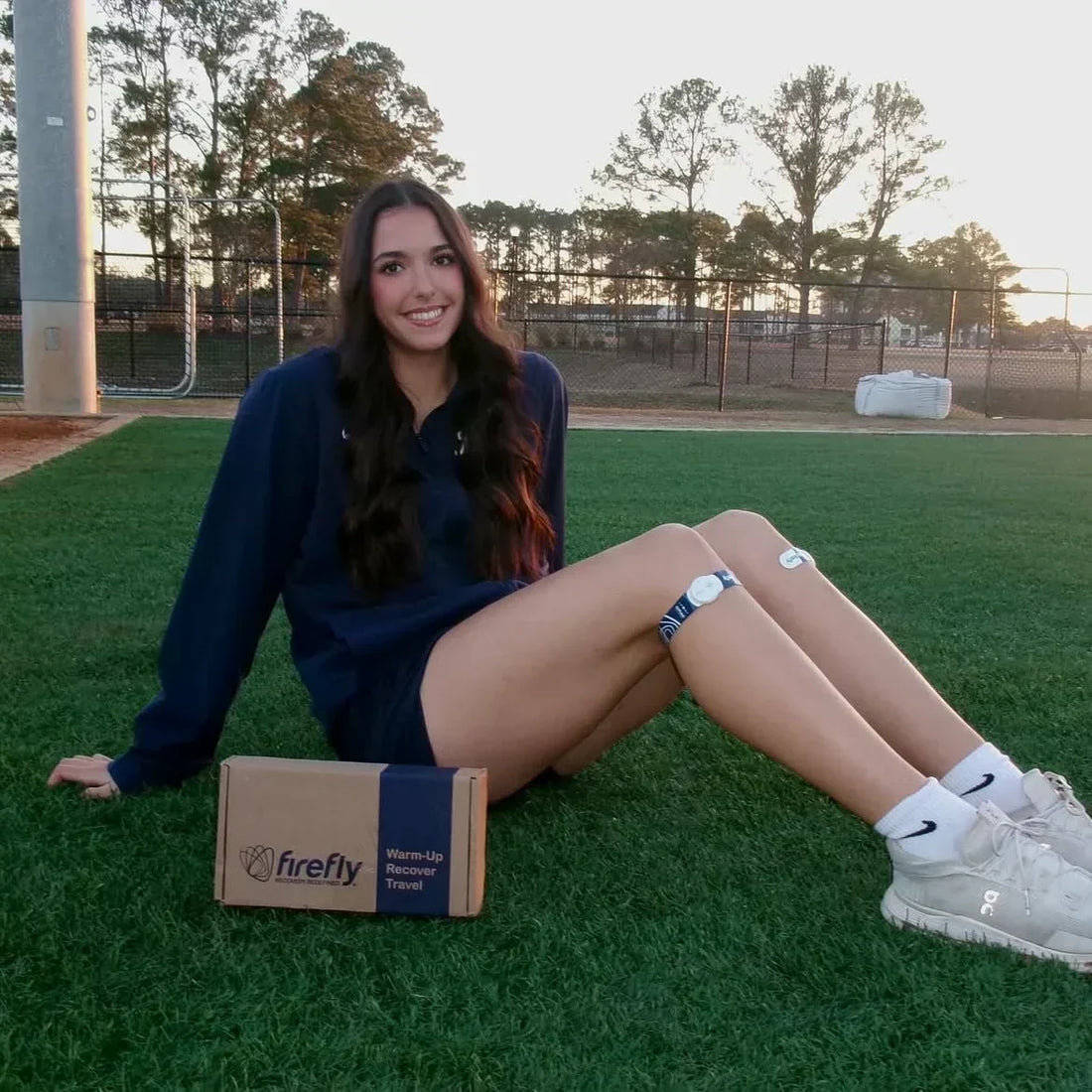 Runner wearing a blue hoodie while using the Firefly Recovery device below the knee after training, seated on a grass turf.